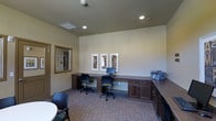 Located at 2770 Copper Peak Lane in Longmont, Colorado, this single-floor business center offers a well-appointed and functional workspace designed to accommodate a range of professional needs. The room features a continuous built-in desk system with rich, dark wood cabinetry that wraps along the walls, providing ample surface area for multiple workstations. Two ergonomic task chairs are positioned at computer terminals, making the space well-suited for productivity. A printer and additional counter space are conveniently integrated into the cabinetry along an adjacent wall, ensuring all essential office equipment is within easy reach. The space is finished with neutral, light-toned walls that create a clean and focused atmosphere, complemented by recessed lighting that provides consistent, even illumination throughout. Decorative framed artwork adds a polished, professional touch to the walls. Patterned carpeting in muted tones grounds the room and contributes to a quiet, comfortable environment. A large framed mirror near the entrance enhances the sense of openness. Overall, this business center is thoughtfully designed to serve as a practical and welcoming workspace.