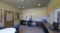 Located at 2770 Copper Peak Lane in Longmont, Colorado, this single-floor business center offers a well-appointed and functional workspace designed to accommodate a range of professional needs. The room features a continuous built-in desk system with rich, dark wood cabinetry that wraps along the walls, providing ample surface area for multiple workstations. Two ergonomic task chairs are positioned at computer terminals, making the space well-suited for productivity. A printer and additional counter space are conveniently integrated into the cabinetry along an adjacent wall, ensuring all essential office equipment is within easy reach. The space is finished with neutral, light-toned walls that create a clean and focused atmosphere, complemented by recessed lighting that provides consistent, even illumination throughout. Decorative framed artwork adds a polished, professional touch to the walls. Patterned carpeting in muted tones grounds the room and contributes to a quiet, comfortable environment. A large framed mirror near the entrance enhances the sense of openness. Overall, this business center is thoughtfully designed to serve as a practical and welcoming workspace.
