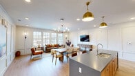 Located at 3389 Endeavor Ln in Vestavia Hills, Alabama, this single-floor open-plan space seamlessly integrates a fully equipped kitchen with a spacious living and dining area. The kitchen serves as a functional centerpiece, anchored by a large island with a grey stone countertop, an undermount sink, and seating for multiple bar-height guests. Warm natural oak cabinetry lines the walls, offering generous storage, while a bold teal accent backsplash adds a distinctive pop of color. Paired brass dome pendant lights illuminate the kitchen with a sophisticated, modern-classic charm. Light hardwood flooring flows continuously throughout the space, enhancing the sense of openness and warmth. The living area is defined by an oversized sofa accented with rust and earth-toned pillows, complemented by leather accent chairs and rounded ottoman ottomans. A multi-arm brass chandelier echoes the kitchen's metallic detailing, tying the two zones together visually. Panel molding on the crisp white walls adds architectural depth and refinement. Large multi-pane windows along the exterior wall flood the space with natural light and offer views of the surrounding neighborhood. A wall-mounted television and sideboard complete the living area, creating a polished, inviting environment ideal for both relaxation and entertaining.