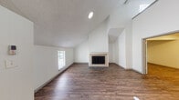 This two-floor home showcases a thoughtfully designed interior with a cohesive aesthetic carried throughout. The living area immediately impresses with its dramatic vaulted ceilings that rise to a striking height, creating an open and airy atmosphere. A central tile-surround fireplace with a classic mantel serves as the focal point of the room, complemented by rich wood-look flooring and neutral white walls that maximize the natural light filtering in through the windows. The adjacent kitchen is well-appointed with grey shaker-style cabinetry, warm stone-toned countertops, under-cabinet lighting, and a full suite of stainless steel appliances including a range, microwave, dishwasher, and refrigerator. A stylish pendant light fixture adds a decorative touch above the dining area. The second floor features additional living or bedroom space with continued wood-look flooring, and a generously sized bathroom complete with a granite-look vanity, grey cabinetry, a large mirror, and a tub-shower combination with a speckled surround and chrome fixtures. A louvered closet door adds practical storage. The textured ceilings and recessed lighting throughout tie the home together with a clean, functional design.