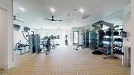 Located at 14811 West Rd in Houston, TX, this fitness center offers a comprehensively equipped exercise facility spread across a single floor. The main exercise room is generously proportioned, featuring stylish multi-tone striped carpet tiles that add a modern aesthetic to the space. Large windows along the perimeter flood the room with natural light and provide views of the surrounding exterior, creating an inviting and energizing atmosphere. Multiple ceiling fans are mounted overhead alongside recessed lighting, ensuring excellent air circulation and consistent illumination throughout. The exercise room is divided into distinct functional zones. A dedicated cardio area houses several treadmills and stationary cycling bikes, each equipped with individual digital displays. Additional cardio options include an elliptical trainer and a rowing machine. The strength training section features a cable and pulley functional trainer, a multi-station weight machine, and an adjustable bench. A well-stocked free weights area includes a full rack of dumbbells, kettlebells of varying weights, and a punching bag for boxing workouts. Floor-to-ceiling mirrors line one wall, enhancing the sense of space and allowing users to monitor their form. A wall-mounted television and a hydration station round out the amenities, making this fitness center a complete and convenient workout destination.