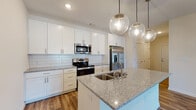 This well-appointed single-floor residence showcases a thoughtfully designed open-concept layout that seamlessly connects the kitchen and living areas. The kitchen is a standout feature, offering crisp white shaker-style cabinetry extending to the ceiling for generous storage, granite countertops with subtle speckled detailing, and a large center island with an integrated sink — ideal for both meal preparation and casual dining. A classic white subway tile backsplash complements the cabinetry, while a suite of stainless steel appliances, including a side-by-side refrigerator with ice and water dispenser, electric range, and over-the-range microwave, elevate the space. Globe-shaped pendant lights hang above the island, adding a contemporary decorative touch. Rich wood-style flooring flows throughout the main living areas, lending warmth and continuity to the space. The living area is spacious and bright, with a ceiling fan and access to natural light through large windows. Bedrooms feature plush carpeting, ceiling fans, and windows that allow in ample natural light. The bathroom is clean and functional, featuring a tiled tub-shower combination with brushed nickel fixtures and wood-style flooring, completing this move-in-ready residence.