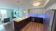 This well-appointed single-floor residence exudes a relaxed coastal aesthetic, combining modern design with comfortable, thoughtfully curated interiors. The open-concept kitchen is a standout feature, boasting dark lower cabinetry contrasted by light upper cabinets, granite countertops, a generous island with bar seating, stainless steel appliances, and striking LED under-cabinet lighting that lends the space a contemporary flair. Rich hardwood-style flooring flows throughout the main living areas.

The living room is bright and inviting, with large sliding glass doors that open to a balcony and frame expansive outdoor views. The space is styled with a sectional sofa, glass coffee table, and coastal-themed artwork, creating an airy, resort-like atmosphere.

The primary bedroom offers plush carpeting, a ceiling fan, walk-in closet, and a spacious en-suite bathroom featuring dual sinks with quartz countertops, a glass-enclosed walk-in shower, and wood-style flooring. A second bedroom carries the coastal theme with soft tones and tasteful beach-inspired décor. An additional bathroom provides a single vanity with quartz countertops and ample storage. Each room benefits from natural light, enhancing the overall bright and welcoming character of this refined coastal retreat.