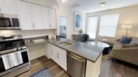 This single-floor residence offers a clean, contemporary design with an open and airy layout. The kitchen serves as the heart of the home, showcasing crisp white shaker-style cabinetry, gray countertops, and a stainless steel dishwasher. A peninsula with an integrated sink extends the workspace and creates a natural division between the kitchen and the adjoining living area, making it ideal for both everyday living and casual entertaining. Wood-look flooring runs throughout, adding warmth and continuity to the space. Large windows fitted with blinds allow for an abundance of natural light while offering privacy, creating a bright and welcoming atmosphere. The open layout provides a direct sightline from the kitchen into the bedroom, which features neutral tones and comfortable furnishings. A decorative accent piece and a small console table add a touch of character near the living area. The overall ambiance is modern and well-appointed, with thoughtful design choices that balance functionality and style throughout the space.