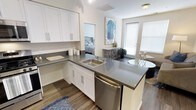 This single-floor residence offers a clean, contemporary design with an open and airy layout. The kitchen serves as the heart of the home, showcasing crisp white shaker-style cabinetry, gray countertops, and a stainless steel dishwasher. A peninsula with an integrated sink extends the workspace and creates a natural division between the kitchen and the adjoining living area, making it ideal for both everyday living and casual entertaining. Wood-look flooring runs throughout, adding warmth and continuity to the space. Large windows fitted with blinds allow for an abundance of natural light while offering privacy, creating a bright and welcoming atmosphere. The open layout provides a direct sightline from the kitchen into the bedroom, which features neutral tones and comfortable furnishings. A decorative accent piece and a small console table add a touch of character near the living area. The overall ambiance is modern and well-appointed, with thoughtful design choices that balance functionality and style throughout the space.