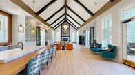 Located in Charlotte, NC, this thoughtfully designed single-floor community space offers a generous and open layout that seamlessly blends architectural character with vibrant, eclectic interiors. The centerpiece of the space is its dramatic vaulted ceiling adorned with bold dark exposed beams, creating an airy, barn-inspired aesthetic that rises impressively overhead throughout the main area.

Light wood-look tile flooring runs throughout, complementing the bright white walls and wainscoting found across the space. The entrance welcomes visitors with striking botanical-print wallpaper, white board-and-batten paneling, and abundant natural light streaming through large windows.

The main living area is anchored by a stunning floor-to-ceiling herringbone-tiled fireplace with a patterned surround, flanked by large windows that offer views of the surrounding landscape. Navy blue barn doors with gold hardware slide open to reveal a secondary living space, offering flexible use for gatherings or quiet lounging.

A kitchen and dining area sit toward the rear of the open floor plan, featuring a marble backsplash, pendant lighting, and communal seating. A vibrant game room with bold orange walls, an arcade machine, and ample window views adds a playful, energetic touch. Together, these areas create a dynamic, welcoming environment ideal for community interaction and relaxation.