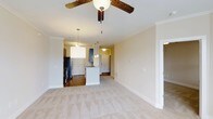 Located at 1000 Antietam Creek Dr in Fayetteville, North Carolina, this single-floor residence offers a thoughtfully designed open-concept layout that seamlessly connects the living and kitchen areas. The living space is finished with neutral-toned carpet and crisp white baseboards, creating a clean and welcoming atmosphere. Crown molding runs along the ceiling, adding a refined architectural touch, while a ceiling fan with wooden blades provides both style and comfort. The kitchen is a standout feature, outfitted with navy blue shaker-style cabinetry, a white kitchen island with pendant lighting above, and stainless steel appliances including a refrigerator and range. Tile flooring in the kitchen area contrasts neatly with the carpeted living space, clearly defining each zone. Pendant lighting and recessed fixtures provide ample illumination throughout. The neutral wall palette of warm beige tones lends a versatile, move-in-ready feel to the entire space. A doorway visible from the main living area hints at additional rooms beyond, suggesting a practical and well-organized floor plan. Overall, the space combines modern finishes with functional design for comfortable single-level living.