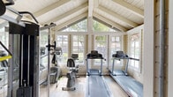 Located at 6506 Doolittle Ave in Riverside, CA, this fitness center is a thoughtfully designed exercise facility housed in a charming, cottage-style building. The main workout area is bright and airy, characterized by a striking vaulted ceiling with exposed white-painted beams that lend an open, spacious feel despite the room's modest footprint. Large multi-pane windows line the walls, flooding the interior with natural light and offering views of the surrounding greenery and an adjacent pool area.

The main space is equipped with cardio machines including treadmills and a recumbent bicycle, as well as a cable weight machine, all arranged to maximize usability. Full-length mirrors along one wall enhance the sense of space and provide functional feedback during workouts. The flooring consists of neutral carpet with green exercise mats placed strategically beneath equipment.

A separate enclosed annex, accessible through a bold orange-framed glass door, provides a more private training area. This secondary room features additional strength and cardio equipment, including an elliptical trainer and a weight bench, set against cool grey walls under standard ceiling lighting. The overall ambiance of the facility is clean, welcoming, and functional, making it a well-equipped community fitness resource.