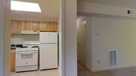 This single-floor space offers a practical and comfortable layout designed for everyday living. The kitchen is a standout feature, illuminated by a skylight that bathes the area in natural light. It is equipped with warm-toned wood upper cabinetry providing ample storage, a white gas range, and a full-size top-freezer refrigerator. A ventilation hood above the range adds functionality to the cooking area. The light-toned flooring in the kitchen complements the neutral wall palette, creating a clean and welcoming atmosphere. Adjacent to the kitchen, a structural column defines the transition into a hallway finished with carpeting that adds warmth underfoot. The hallway leads to additional rooms, with a door visible at the far end suggesting a private space such as a bedroom or storage area. A wall-mounted thermostat is conveniently located in the hallway, indicating climate control throughout the unit. The overall design is straightforward and efficient, with neutral tones and practical finishes that create a cohesive, move-in-ready environment suited for comfortable day-to-day living.