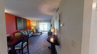 This single-floor residence is thoughtfully designed with a bold, energetic aesthetic and functional layout. The open living and dining area serves as the heart of the home, featuring plush carpeted flooring and striking accent walls in deep orange and chartreuse green that create a warm, lively atmosphere. A large sliding glass door floods the space with natural light and provides direct access to a private patio with outdoor views. A corner entryway table and textured ceiling add character throughout.

The kitchen is efficiently arranged with dark espresso cabinetry, granite-look countertops, black appliances including a range and dishwasher, and a drop-panel ceiling with integrated lighting. Wood-look flooring grounds the space with a clean, modern contrast to the cabinetry.

Two well-proportioned bedrooms each feature carpet, individual accent walls—one in teal, another in slate gray—and ample natural light from windows with curtains. The primary bedroom includes a private en-suite bathroom with a tub-shower combination, a large framed mirror, and a navy vanity. A second full bathroom features dark wood-look flooring, a tub-shower combination, and clean white finishes, providing practical functionality for residents and guests alike.