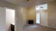 This two-story home showcases a spacious main living area distinguished by soaring vaulted ceilings that amplify the sense of openness and airiness throughout the space. A ceiling fan is mounted high above, serving both as a functional element and a visual anchor for the room. Natural light filters in through blinded windows, lending a bright and welcoming atmosphere to the interior.

A corner brick fireplace serves as the living area's focal point, adding a classic, traditional character to the room. It sits on a tiled hearth that contrasts with the surrounding wall-to-wall carpeting, which extends throughout the main living space in a neutral tone that complements the warm beige wall color.

Adjacent to the living area, a bathroom is visible, featuring cabinetry with a dark wood finish and a sink, offering convenient access from the main floor. The overall layout suggests a thoughtful floor plan that balances communal and private spaces across its two levels. The neutral color palette, combined with the architectural details of the vaulted ceiling and fireplace, creates a comfortable and inviting environment throughout the home.