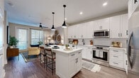 This 1-bedroom, 1-bathroom residence offers a thoughtfully designed single-floor layout that maximizes every square foot. The open-concept kitchen and living area serves as the heart of the home, anchored by a striking white shaker-style kitchen featuring floor-to-ceiling cabinetry, a white subway tile backsplash, stainless steel appliances including a french door refrigerator, a gas range, and a built-in microwave. A central island with a white countertop and pendant lighting overhead provides both additional prep space and casual seating for two. Wood-look flooring runs throughout, lending warmth and continuity to the space. The adjoining living room is bright and inviting, with large windows and a glass door allowing natural light to fill the room, complemented by a ceiling fan overhead. The bedroom features a bold teal accent wall, creating a calm and stylish retreat with ample room for a full-size bed and additional furnishings. The bathroom offers a dark-toned vanity with a white countertop, a large mirror, a tub-shower combination, and direct access to a walk-in closet with built-in shelving, adding practical storage to the unit's overall appeal.