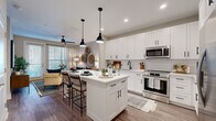 This 1-bedroom, 1-bathroom residence offers a thoughtfully designed single-floor layout that maximizes every square foot. The open-concept kitchen and living area serves as the heart of the home, anchored by a striking white shaker-style kitchen featuring floor-to-ceiling cabinetry, a white subway tile backsplash, stainless steel appliances including a french door refrigerator, a gas range, and a built-in microwave. A central island with a white countertop and pendant lighting overhead provides both additional prep space and casual seating for two. Wood-look flooring runs throughout, lending warmth and continuity to the space. The adjoining living room is bright and inviting, with large windows and a glass door allowing natural light to fill the room, complemented by a ceiling fan overhead. The bedroom features a bold teal accent wall, creating a calm and stylish retreat with ample room for a full-size bed and additional furnishings. The bathroom offers a dark-toned vanity with a white countertop, a large mirror, a tub-shower combination, and direct access to a walk-in closet with built-in shelving, adding practical storage to the unit's overall appeal.