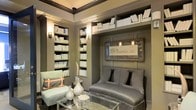 This single-floor space exudes refined sophistication, centered around a beautifully appointed library lounge that serves as its defining feature. The room is dressed in a harmonious palette of muted greens and warm grays, lending an air of understated elegance throughout. Floor-to-ceiling built-in bookshelves line the walls, filled with curated volumes that reinforce the scholarly, cultivated character of the space. Decorative wall sconces are nestled within the shelving, casting a warm, ambient glow that enhances the intimate atmosphere. A tufted gray settee is positioned against a paneled wall beneath a gilded framed artwork, creating a distinguished focal point. Complementing it is a light upholstered accent chair adorned with a patterned throw pillow, adding a subtle contrast in texture and tone. A sleek glass-topped coffee table anchors the seating arrangement, styled with sculptural vases and decorative objects that add visual depth. Crown molding frames the ceiling, elevating the architectural detail and reinforcing the space's classic sensibility. The overall ambiance is one of quiet luxury — a thoughtfully designed retreat that balances function and aesthetics with a polished, gallery-like quality.