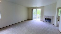 This single-floor residence offers a clean, understated design with a welcoming and functional layout. The living area serves as the heart of the space, featuring wall-to-wall carpeting that adds warmth and comfort underfoot. Neutral-toned walls and a simple, open layout create a bright and airy ambiance throughout.

A standout feature of the living area is the fireplace, positioned along one wall, providing a cozy focal point for relaxation. Adjacent to the fireplace, large sliding glass doors flood the room with natural light and open directly onto a private balcony. The balcony is framed by a white railing and overlooks a lush, tree-lined outdoor setting, offering a serene and green view that enhances the overall atmosphere of the space.

The sliding doors allow for a seamless indoor-outdoor connection, making the balcony an inviting extension of the living area. The ceiling is smooth and uniform, contributing to the clean, uncluttered aesthetic of the interior. Overall, this space presents a practical and comfortable layout that balances simplicity with key lifestyle features, including natural light, outdoor access, and a fireplace — making it a well-rounded and inviting place to call home.