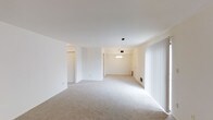 This single-floor space offers a clean, open layout designed for comfortable everyday living. The interior is finished in a neutral white and cream palette throughout, creating a bright and airy atmosphere that feels welcoming and versatile.

The main living area is generously proportioned, featuring soft, plush wall-to-wall carpeting in a light beige tone that adds warmth underfoot. Large vertical blinds line the windows, allowing for adjustable natural light and contributing to the room's open, expansive feel. A ceiling-mounted HVAC vent ensures year-round comfort.

Flowing seamlessly from the living area is a dedicated dining space, distinguished by a classic chandelier that serves as a subtle focal point. The dining area features paneled wall detailing, adding a touch of traditional character to the otherwise minimalist interior. A floor-mounted baseboard heating unit is positioned along the wall, providing practical climate control.

A hallway visible from the main living area leads to additional rooms, with a coat closet or storage space nearby, contributing to the home's functional layout. The consistent flooring, neutral wall tones, and connected flow between spaces make this a cohesive and easy-to-navigate single-level home well-suited for a variety of furnishing and lifestyle needs.