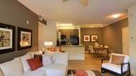This single-floor residence offers a thoughtfully designed open-concept layout that seamlessly connects the living, dining, and kitchen areas into one cohesive living environment. The living room is generously sized, anchored by a plush sectional sofa accented with warm red and neutral throw pillows, and finished with light hardwood flooring that flows throughout the space. A ceiling fan overhead adds both style and function. The kitchen features dark cabinetry paired with granite countertops, black appliances including a refrigerator, microwave, and stove, and a peninsula that serves as both a prep space and a casual breakfast bar. Track lighting illuminates the kitchen and dining area effectively. The dining area is positioned adjacent to the kitchen, offering a rectangular wooden table with modern white chairs, ideal for everyday meals or entertaining. Two framed art pieces in red and neutral tones adorn the accent wall, adding a pop of color that ties the room together. The warm earth-tone color palette — featuring deep brown accent walls contrasted with bright white trim and ceilings — gives the space a modern yet inviting ambiance throughout.