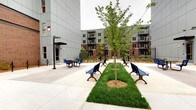 This well-appointed residential community combines contemporary architecture with thoughtfully designed communal amenities. The exterior presents a striking mix of classic red brick and modern grey paneling across multiple stories, with private balconies overlooking the grounds. At the heart of the community lies a generous landscaped courtyard, featuring a lush open lawn framed by manicured plantings and winding pathways — an ideal setting for relaxation and casual gatherings. An adjacent hardscaped plaza offers additional outdoor seating, with blue benches arranged around young shade trees, creating an inviting social hub for residents. Inside, the community includes a dedicated dog wash facility, thoughtfully finished with clean white subway tile wainscoting, polished concrete floors, and large black-framed windows that bring in natural light. The space is equipped with a stainless steel grooming tub complete with a spray hose and non-slip entry ramp, making pet care convenient and accessible. Colorful canine-themed artwork adorns the walls, adding a playful touch to the functional space. Together, these features reflect a community designed with both residents and their pets in mind, blending practical amenities with a welcoming and modern aesthetic.