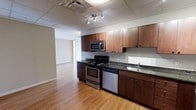 This single-floor space offers a functional and modern layout designed for comfortable everyday living. The kitchen serves as a central highlight, featuring rich dark wood cabinetry with sleek hardware, dark granite countertops, and a full suite of stainless steel appliances including an electric range, built-in microwave, and dishwasher. A double-basin sink is positioned along the counter for added convenience. The kitchen is illuminated by a contemporary track lighting fixture mounted to a drop ceiling with integrated ventilation, ensuring a bright and practical workspace. Warm-toned hardwood flooring runs throughout, adding a sense of continuity and warmth to the entire space. The kitchen flows openly into an adjacent living area, which benefits from natural light and a neutral color palette of soft whites and warm beige tones on the walls. The open-concept design creates a spacious and airy feel, making the space feel larger and more connected. Clean lines and a well-considered layout make this a move-in-ready space suited to a variety of lifestyles.
