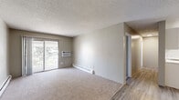 Located at 1869 Coronado Pkwy N in Denver, Colorado, this single-floor residence offers three bedrooms and one bathroom within a efficiently arranged floor plan. The combined kitchen and living area serves as the heart of the home, where light hardwood-style flooring runs through the kitchen, which is equipped with white cabinetry, a double-basin sink, white appliances including a range, microwave, and refrigerator, and a skylight-style ceiling panel paired with a ceiling fan. The living room extends from this space, featuring plush carpet, a wall-mounted air conditioning unit, baseboard heating, and a sliding glass door that opens to a balcony with views of mature trees, allowing natural light to fill the room. A textured popcorn ceiling is consistent throughout the residence. Each of the three bedrooms is carpeted and includes a built-in closet with sliding or hinged doors and rod storage. The bedrooms share a full bathroom appointed with a tub-shower combination, toilet, and vanity. Neutral, warm-toned walls throughout provide a clean and versatile backdrop. Baseboard heating is present in multiple rooms, ensuring comfort year-round. The entrance transitions seamlessly between the hardwood-style and carpeted areas of the home.
