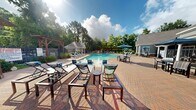 Located at 14600 Huffmeister Rd in Cypress, TX 77429, this resort-style outdoor pool area offers a spacious and well-appointed recreational retreat for residents. The centerpiece is a large, freeform swimming pool with crystal-clear turquoise water, designed to accommodate both relaxed leisure and active swimming. The pool deck is finished in warm brick-toned pavers and is lined with multiple blue chaise lounge chairs, providing ample space for sunbathing and relaxation. A prominent wooden pergola structure anchors one side of the deck, offering shaded seating beneath its open-beam canopy. The pool features built-in water elements, including in-pool lounge chairs with gentle water jets, adding a resort-like ambiance to the experience. An adjacent single-story clubhouse building with a pitched roof, arched window detail, and a covered outdoor patio with umbrella-shaded seating serves as a functional hub for the pool area. A smaller brick accessory structure, likely housing pool equipment or restrooms, is also present on the grounds. The entire space is enveloped by tall, mature trees that provide natural privacy, shade, and a lush green backdrop. Open skies above allow for abundant natural light throughout the day, enhancing the overall outdoor atmosphere of this well-maintained community amenity.