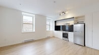 This studio on Rue Sherbrooke Ouest in Montréal offers a well-designed, single-floor layout that makes efficient use of its compact footprint. The open-concept main area combines living and kitchen functions in one bright, airy space. Large double-hung windows flood the room with natural light and offer views of the surrounding urban neighbourhood, contributing to an open and welcoming ambiance.

The kitchen is thoughtfully integrated along one wall, featuring sleek white cabinetry, a bold black tile backsplash, stainless steel appliances including a full-size range and refrigerator, and a built-in range hood — a clean contrast of modern and functional design. Light hardwood flooring runs throughout the main living area, adding warmth and continuity to the space. A wall-mounted mini-split unit provides climate control.

A hallway connects the main space to the bathroom, which is finished with classic white square tile covering both the walls and floor. The bathroom includes a glass-enclosed shower stall, a toilet, and a vanity, all maintained in a crisp, clean aesthetic.

With its modern finishes, natural light, and practical layout, this studio offers a functional and contemporary urban living space in a prime Montréal location.