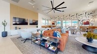 Located at 32000 N 29th Ave in Phoenix, AZ, this contemporary single-floor community space offers a thoughtfully designed environment spanning a generous footprint across three distinct areas. The living room serves as the heart of the space, showcasing an open-concept layout with soaring ceilings, recessed lighting, and an oversized statement ceiling fan. Bold design choices abound, including an artistic botanical mural accent wall, large floor-to-ceiling windows that flood the interior with abundant natural light, and direct views of an outdoor patio area with shaded seating. Comfortable lounge furniture in warm terracotta and cool aqua tones is complemented by a built-in media wall and casual banquette seating along the windowed perimeter. A sleek kitchen bar with cobalt blue stools and a modern multi-bulb chandelier anchors an adjacent gathering area. The hallway entry features clean white walls, decorative greenery, black-framed glass doors, and curated wall art, creating an inviting transition into the space. The private office is distinguished by a sage-green accent wall, a polished marble-top worktable, sculptural globe pendant lighting, and ergonomic seating — ideal for focused work. Light tile flooring and a neutral palette unify the space with a bright, modern Arizona aesthetic.