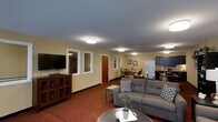 Located in Rolesville, this single-floor space offers a well-organized and functional layout that integrates living, dining, and kitchen areas into a cohesive, open floor plan. The living area is anchored by a plush gray sofa accented with decorative pillows, arranged around a central coffee table and complemented by a media console with a wall-mounted flat-screen television. Flush-mount ceiling lights provide consistent, even illumination throughout the space. Warm yellow-toned walls contribute to an inviting and comfortable ambiance, while rich red carpeting adds a bold, grounding element underfoot. A transition into the dining area reveals a classic wooden dining set, ideal for gatherings and daily meals. The kitchen, visible in the background, is equipped with dark cabinetry, a white refrigerator with a water dispenser, and dark countertops, offering practical utility without sacrificing style. Interior windows and a wood-toned door along one wall add architectural character and allow light to flow between areas. Framed artwork on the walls and a small potted plant on the coffee table introduce subtle decorative touches that enhance the overall warmth of the space. The thoughtful arrangement of furnishings and fixtures makes this a practical and welcoming environment.