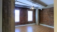 This single-floor loft space showcases a compelling blend of industrial heritage and modern comfort. The standout feature is the full-height exposed brick wall, rich in texture and color, which lends an authentic, historic character to the interior. Structural concrete columns and heavy timber ceiling beams run throughout, underscoring the building's original construction and adding a raw, architectural appeal. The hardwood flooring provides a warm contrast to the rugged masonry, creating a balanced and inviting atmosphere. Natural light floods the space through multiple large windows fitted with blinds, each framing bright, airy views while illuminating the interior with a soft, diffused glow. A ceiling fan with an integrated light fixture is centrally mounted, ensuring both air circulation and ambient lighting year-round. The open layout offers exceptional flexibility, making the space well-suited for a variety of uses. Overhead, industrial-style tracking hardware is visible along the ceiling, adding to the loft's utilitarian aesthetic. The overall ambiance is one of refined industrial character — a space that honors its architectural past while remaining highly functional and adaptable.