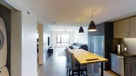 This single-floor space showcases a thoughtfully designed open-concept layout that integrates the kitchen, dining, and living areas into one cohesive environment. The kitchen is equipped with dark cabinetry reaching toward the ceiling, a stainless steel refrigerator, and is anchored by a butcher block island that doubles as a dining surface, surrounded by woven accent chairs. Two matte black pendant lights hang above the island, providing focused task lighting while adding a modern design element. Dark hardwood-style flooring runs throughout, creating a strong contrast against the white walls and ceiling. The living area offers a comfortable sectional sofa arranged around a flat-screen television mounted on the wall, with a light-toned area rug defining the space. A ceiling fan with integrated lighting serves the living area, which also benefits from a large window that invites natural light and provides an outdoor view, contributing to an open and airy atmosphere. A hallway visible along one side suggests additional rooms beyond the main living space. Overall, the design balances functionality with a clean, contemporary aesthetic.