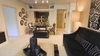 This single-floor residence showcases a cohesive open-concept design that connects the living, dining, and kitchen areas in one flowing layout. The living area is anchored by a sleek black sofa accented with decorative pillows, complemented by a textured area rug and a contemporary coffee table styled with curated decorative objects. A striking black leather ottoman serves as a functional centerpiece between the living and dining spaces.

The dining area features a dark, reflective table surrounded by elegant houndstooth-upholstered chairs, with silver decorative accents adding a refined touch. A gallery-style arrangement of circular framed pieces adorns one wall, contributing to the space's bold aesthetic.

The kitchen is visible through a structural opening, revealing dark cabinetry, stainless steel appliances including a built-in microwave, and light countertops that contrast the cabinetry. Track lighting runs along the ceiling throughout, ensuring consistent illumination across all areas. A hallway leading toward additional rooms and an entry door is visible in the background, suggesting a well-organized and functional floor plan. The overall ambiance is sophisticated, with a consistent black, white, and neutral color palette throughout.