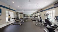 This fitness center is designed to support a comprehensive workout experience within a generously proportioned, single-floor space. The layout is open and well-organized, with dark rubber flooring throughout that provides both durability and traction during exercise. A distinctive teal accent stripe runs along the upper portion of the cream-colored walls, lending the space a clean, professional aesthetic.

The cardio zone features multiple treadmills and elliptical machines, each equipped with individual display consoles. Several wall-mounted flat-screen televisions are positioned for easy viewing from the cardio equipment. Natural light flows in through multiple large windows, including an arched window at one end, creating a bright and energizing atmosphere.

The strength training area is equally well-appointed, offering a multi-station cable machine with various attachments, a squat rack with a barbell and weight plates, and a dedicated free weights section with a full rack of dumbbells. Adjustable workout benches are available throughout. Full-length mirrors line the walls, visually expanding the space and allowing users to monitor their form. Recessed lighting supplements the natural light, ensuring the entire facility is consistently well-illuminated. The textured ceiling and clean finishes complete the polished, functional character of this fitness facility.