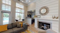 This thoughtfully designed single-floor clubhouse offers an inviting and stylish environment for residents to gather and relax. The main lounge area commands immediate attention with its soaring ceilings, floor-to-ceiling windows, and transom windows that flood the space with abundant natural light. Large glass doors provide direct views of the outdoor pool area, seamlessly connecting the interior to the exterior. Light hardwood flooring runs throughout, complementing the neutral, warm color palette.

A dramatic shiplap accent wall serves as the focal point of the lounge, framing a sleek fireplace beneath a decorative round mirror. The seating area features comfortable sofas and swivel chairs arranged around a patterned area rug with gold and grey tones, creating an inviting social atmosphere. A wall-mounted flat-screen television adds a functional entertainment element to the space.

Adjacent to the lounge, a kitchenette with a white peninsula counter, pendant lighting, and dark cabinetry provides convenient amenity access. A dedicated private office with natural light offers a quiet workspace. Decorative touches throughout — including bold abstract artwork, an array of gold sunburst wall décor, and curated accessories — give the clubhouse a polished, contemporary aesthetic.