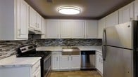 This single-floor space showcases a thoughtfully designed kitchen that serves as its standout feature. The kitchen is outfitted with crisp white cabinetry extending from floor to ceiling, providing ample storage while contributing to a clean, bright aesthetic. A eye-catching mosaic tile backsplash in muted tones of gray, black, and beige adds texture and visual interest, elegantly bridging the upper and lower cabinetry. The countertops feature a marble-style surface that lends a refined, upscale feel to the space. Stainless steel appliances, including a gas range, a built-in dishwasher, and a full-size refrigerator, provide a modern, cohesive look. A deep stainless steel farmhouse sink serves as a functional centerpiece beneath the upper cabinets. Overhead, a large flush-mount ceiling light provides even, ambient illumination throughout the kitchen. The flooring features a neutral tile that complements the overall color palette. The textured ceiling adds a classic touch, while the dark-toned upper walls create a cozy contrast against the bright white cabinetry. Overall, the space exudes a well-balanced combination of modern updates and warm, inviting design elements.