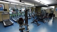 This fitness center offers a comprehensive workout environment designed to accommodate a variety of exercise routines. The space is lined with floor-to-ceiling windows along one wall, flooding the interior with natural light and providing views of lush outdoor greenery, creating an energizing atmosphere for exercise. Mirrored walls further enhance the sense of openness and allow users to monitor their form during workouts.

The facility is well-stocked with a strong selection of cardio equipment, including multiple treadmills and elliptical machines arranged in a row along the windowed wall. Stationary and upright cycling bikes are also available for low-impact cardio training. A cable resistance machine positioned prominently in the foreground provides options for targeted strength and functional training exercises.

Overhead, ceiling fans are mounted to aid in air circulation throughout the workout area. The flooring is a durable blue rubberized surface, practical and supportive for high-impact activity. Framed artwork on the walls adds a touch of polish to the utilitarian setting. Overall, the space strikes a balance between functionality and comfort, making it a practical and inviting fitness facility.