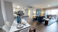 This well-appointed single-floor residence offers an open-concept layout that effortlessly connects the dining area, kitchen, and living room into one cohesive, functional living space. Rich hardwood-style flooring flows throughout, anchoring the interior with warmth and a sense of continuity.

The dining area is highlighted by a sleek marble-top table, elegantly styled with decorative place settings and a striking blue floral centerpiece that adds a pop of color. Adjacent to the dining space, the kitchen features a dark-topped breakfast bar that provides additional seating and workspace, complemented by white cabinetry visible in the background.

The living area is tastefully furnished with deep teal accent chairs, a well-curated area rug, and a large statement artwork that commands the main wall. Warm ambient lighting from carefully placed lamps creates a cozy and inviting atmosphere throughout the space.

Neutral gray walls provide a versatile backdrop that complements the bold accents of teal and gold woven throughout the decor. Recessed lighting and a skylight contribute to a bright, airy environment. A hallway visible beyond the kitchen leads to additional private areas of the residence, suggesting a thoughtful and practical floor plan designed for comfortable everyday living.