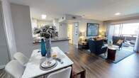 This well-appointed single-floor residence offers an open-concept layout that effortlessly connects the dining area, kitchen, and living room into one cohesive, functional living space. Rich hardwood-style flooring flows throughout, anchoring the interior with warmth and a sense of continuity.

The dining area is highlighted by a sleek marble-top table, elegantly styled with decorative place settings and a striking blue floral centerpiece that adds a pop of color. Adjacent to the dining space, the kitchen features a dark-topped breakfast bar that provides additional seating and workspace, complemented by white cabinetry visible in the background.

The living area is tastefully furnished with deep teal accent chairs, a well-curated area rug, and a large statement artwork that commands the main wall. Warm ambient lighting from carefully placed lamps creates a cozy and inviting atmosphere throughout the space.

Neutral gray walls provide a versatile backdrop that complements the bold accents of teal and gold woven throughout the decor. Recessed lighting and a skylight contribute to a bright, airy environment. A hallway visible beyond the kitchen leads to additional private areas of the residence, suggesting a thoughtful and practical floor plan designed for comfortable everyday living.