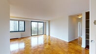 This well-organized residential unit spans a single floor and is defined by its gleaming parquet wood flooring, which extends consistently throughout the living spaces and bedrooms, adding warmth and character to the interior. The spacious living and dining area is a standout feature, bathed in natural light through large sliding glass doors that open onto a private balcony overlooking a lush urban skyline. A side window further enhances the brightness of this generous main living space. The kitchen is clean and functional, featuring gray laminate countertops, a gas cooktop, white cabinetry, and classic black-and-white diamond-patterned tile flooring. The bedrooms are well-proportioned, each offering natural light through large windows with urban views, and are equipped with built-in closets featuring bifold doors and shelving. The primary bedroom benefits from an en-suite bathroom tiled in gray, complete with a bathtub. A second bathroom features retro mint-green tile and a matching tub, adding a charming vintage touch. Radiator heating units are present throughout, and the neutral white walls provide a versatile backdrop. The overall layout, as seen in the dollhouse view, is practical and spacious, making excellent use of the available floor plan.