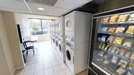 This single-floor communal laundry facility is designed for practicality and convenience. The room is lined with multiple stacked washer and dryer units arranged in a neat row along one wall, maximizing capacity within the space. The machines are modern front-loading models, offering efficient operation for residents or guests. Light ceramic tile flooring runs throughout, providing a clean and easy-to-maintain surface. Natural light filters in through a large window along one wall, brightening the interior and creating a pleasant atmosphere. Recessed ceiling lighting supplements the natural light, ensuring the space remains well-lit at all times. A snack and beverage vending machine is conveniently positioned near the entrance, adding an extra layer of amenity for users during their wait. A small seating area with a table and chairs is tucked near the window, offering a comfortable spot to sit while laundry cycles complete. The drop ceiling with standard panels contributes to the room's functional, no-frills aesthetic, keeping the focus on utility and ease of use.