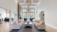 This single-floor clubhouse is an elegantly designed gathering space that seamlessly blends sophisticated architectural details with comfortable, functional amenities. The grand main living area commands attention with its soaring coffered ceilings, an ornate multi-arm chandelier, and a full-height stacked stone fireplace that serves as a striking focal point. Two generous sofas flank a central coffee table, while a wall of tall multi-pane windows floods the room with natural light and frames picturesque views of the outdoor pool and landscaped grounds. The impressive entrance foyer features a dramatic arched ceiling with a lantern pendant, setting a welcoming tone from the moment of arrival. Arched doorways and hallways connect the various areas, maintaining an open, flowing layout throughout. The fully equipped kitchen offers white cabinetry, dark granite countertops, stainless steel appliances, and a tiled peninsula with bar seating, complemented by stylish industrial pendant lighting. A dedicated game room with a pool table and pool views provides a relaxed recreational retreat, while a separate lounge area with tufted seating and display shelving offers a quieter setting for conversation. Warm neutral tones, wainscoting, crown molding, and tile flooring throughout unify the space with a refined yet inviting character.