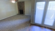 This single-floor residence offers a comfortable and functional layout designed for everyday living. The main living area is a welcoming space highlighted by a striking floor-to-ceiling stone corner fireplace, which serves as a natural focal point and adds a rustic, warm character to the room. Neutral-toned carpet covers the main living area, providing a soft and cozy feel underfoot, while hardwood flooring near the entryway adds a classic, durable contrast. A ceiling fan is centrally positioned overhead, offering both practical airflow and understated style. Natural light enters the space through a full-length glass front door fitted with blinds, brightening the entryway and creating a welcoming transition between the interior and exterior. The walls are finished in a soft, neutral tone that complements the earthy textures of the stone fireplace and flooring materials. The overall ambiance is casual and comfortable, with a layout that balances openness with warmth. The combination of natural materials — stone, wood, and carpet — gives this home a grounded, livable character well-suited to a variety of furnishing styles.