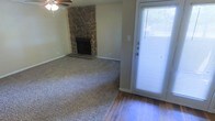 This single-floor residence offers a comfortable and functional layout designed for everyday living. The main living area is a welcoming space highlighted by a striking floor-to-ceiling stone corner fireplace, which serves as a natural focal point and adds a rustic, warm character to the room. Neutral-toned carpet covers the main living area, providing a soft and cozy feel underfoot, while hardwood flooring near the entryway adds a classic, durable contrast. A ceiling fan is centrally positioned overhead, offering both practical airflow and understated style. Natural light enters the space through a full-length glass front door fitted with blinds, brightening the entryway and creating a welcoming transition between the interior and exterior. The walls are finished in a soft, neutral tone that complements the earthy textures of the stone fireplace and flooring materials. The overall ambiance is casual and comfortable, with a layout that balances openness with warmth. The combination of natural materials — stone, wood, and carpet — gives this home a grounded, livable character well-suited to a variety of furnishing styles.