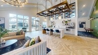 This thoughtfully designed single-floor community clubhouse offers a generous and versatile layout finished with upscale architectural details throughout. The centerpiece of the space is a grand open-concept lounge and social area, featuring soaring vaulted ceilings, a striking geometric metal overhead installation with pendant lighting, and expansive herringbone-patterned flooring that flows seamlessly across the space. Floor-to-ceiling glass doors flood the area with natural light and provide views of the surrounding outdoor spaces. A sleek white kitchenette island with navy bar seating and open shelving serves as a functional social hub. The living room area exudes warmth with a whitewashed brick accent wall, a modern linear fireplace, a mounted television, and comfortable lounge seating. An adjacent recreation area features soaring ceilings with exposed beams, large windows, and a billiards table set atop a bold patterned carpet, ideal for leisure and entertainment. The private office is a refined, well-lit conference room outfitted with a solid wood table, upholstered seating, a wall-mounted display, and a textured area rug. Wide hallways adorned with large-scale artwork and vaulted ceilings connect each area, reinforcing the space's cohesive modern aesthetic.