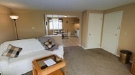 This single-floor space offers a practical and comfortable open-concept layout that seamlessly connects the living, dining, and kitchen areas. The living area is carpeted and centered around a plush sofa accented with patterned throw pillows and a blanket, complemented by a warm wood-and-glass coffee table — creating an inviting, relaxed atmosphere. A half-wall partition with a pass-through window separates the living space from the dining and kitchen areas, adding a sense of definition without compromising the open feel. The dining area features a classic round table with elegant chairs, positioned near a window that allows natural light to fill the space. The kitchen is efficiently laid out with white appliances, including a range and refrigerator, and upper cabinetry that provides ample storage. The warm beige tones carried throughout the walls and flooring create a cohesive and neutral palette. A private door on one side of the living area leads to additional space, adding versatility to the layout. Overall, this space balances functionality and comfort, making it well-suited for everyday living.