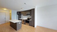 This single-floor space showcases a contemporary open-concept layout centered around a well-appointed kitchen. The kitchen is outfitted with a full suite of stainless steel appliances, including a refrigerator, electric range, built-in microwave, and dishwasher. Dark espresso-toned cabinetry extends from counter height to the ceiling, providing generous storage while creating a bold visual contrast against the crisp white walls and light countertops. A central kitchen island with a matching dark finish offers additional workspace and casual seating potential. Light-toned hardwood flooring runs throughout, lending warmth and continuity to the space. Recessed track lighting above the kitchen provides focused illumination, complementing the natural light that fills the open area. The seamless transition between the kitchen and the adjoining living area creates a spacious, airy feel ideal for both everyday living and entertaining. Clean lines, neutral tones, and modern finishes define the overall aesthetic, giving the space a polished and functional character.