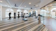 Located at 2470 Merchant Street in Frederick, Maryland, this single-floor fitness center combines elegant architectural design with a comprehensive array of exercise amenities. The main workout area is generously proportioned, featuring distinctive striped carpeting, coffered ceilings with recessed lighting, and multiple ceiling fans that maintain a comfortable atmosphere. Tall arched windows with white blinds allow ample natural light to fill the space, creating an open and inviting environment. The main floor is equipped with a full lineup of cardio machines — including treadmills, ellipticals, and stationary bikes — as well as a cable weight machine, free weights, and adjustable benches, accommodating a wide range of fitness routines. A wall-mounted television and built-in shelving add functionality to the space. A separate studio room, enclosed by elegant etched-glass partition doors, provides a dedicated area for flexibility and functional training. This room features hardwood flooring and is stocked with stability balls, medicine balls, and a dumbbell rack. The entryway connecting the two areas is finished with warm hardwood floors, crown molding, and crisp white wainscoting, reflecting the refined aesthetic that defines the facility throughout.