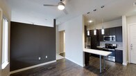 This single-floor space showcases a contemporary open-concept design that seamlessly connects the living area and kitchen. The living space is defined by rich, dark hardwood flooring that extends throughout, complemented by crisp white walls and a striking dark charcoal accent wall that adds depth and visual contrast. A ceiling fan with an integrated light fixture is centrally positioned, providing both airflow and ambient lighting.

The kitchen is thoughtfully appointed with dark espresso cabinetry reaching toward the ceiling, stainless steel appliances including a refrigerator and microwave, and a clean white countertop that wraps around to form a breakfast bar with sleek metal bar stools. Three modern cylindrical pendant lights hang above the counter, offering focused task lighting while enhancing the contemporary aesthetic. Recessed ceiling lights provide additional illumination throughout the kitchen area.

A hallway visible in the background suggests access to additional rooms, and the transition from dark hardwood to lighter flooring in that corridor hints at distinct zones within the space. Overall, the unit carries a clean, modern ambiance with a cohesive palette of dark and light tones.