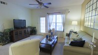 This single-story residence offers a welcoming and well-organized layout designed for comfortable everyday living. The living room serves as the central gathering space, featuring light-colored walls, plush carpeting, and an overhead ceiling fan that enhances air circulation throughout the room. A glass-topped coffee table with a lower shelf anchors the seating arrangement, which includes cream-toned sofas and chairs accented with navy blue decorative pillows, lending a cohesive and tasteful aesthetic. Natural light fills the room through multiple windows dressed with sheer curtains and decorative drapes, creating a bright and airy atmosphere. A glass-panel door provides additional access and light. A media console with glass-front cabinets is positioned along one wall, offering functional storage.

The space transitions naturally into a dedicated dining area, distinguished by a warm golden-yellow accent wall that adds depth and visual interest. A round dining table surrounded by dark wood chairs sits beneath a pendant light fixture, creating an intimate setting for meals. The open sightlines between the living and dining areas promote a sense of spaciousness and connectivity throughout the home.