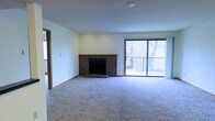This single-floor space presents a spacious, open living area defined by clean lines and neutral tones. The room is anchored by a classic brick fireplace, complete with a raised brick hearth and a simple wood mantel, serving as a natural focal point. Its dark brick construction adds a traditional, grounded character to the otherwise light and airy interior.

Soft, neutral carpeting extends throughout the living space, contributing to a warm and comfortable atmosphere. The walls are finished in a clean off-white tone, complementing the understated aesthetic and allowing for versatile furnishing options.

A large sliding glass door floods the room with natural light, creating a bright and welcoming environment. The door opens to a private balcony enclosed by a metal railing, offering an outdoor extension of the living space with views of surrounding trees, adding a touch of natural scenery. The combination of the fireplace and the outdoor access makes this space well-suited for both relaxed everyday living and entertaining. A doorway visible along one wall suggests additional rooms beyond, completing this well-appointed single-floor layout.