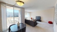 This single-floor residence offers a thoughtfully arranged layout that balances comfort and functionality. The open living area serves as the heart of the home, featuring plush carpeting, a large sliding glass door that opens to a private balcony with outdoor views, and a wide window fitted with vertical blinds that fills the room with natural light. The space comfortably accommodates a full seating arrangement, making it ideal for both relaxing and entertaining. A dedicated dining area with a pendant light fixture sits adjacent to the living room, providing a seamless flow throughout the common areas. A distinctive abstract artwork adorns the wall, adding a touch of character to the neutral, warm-toned palette found throughout.

The galley-style kitchen is a standout feature, finished with a bold black-and-white checkered tile floor, light wood cabinetry, white countertops, and stainless steel appliances including a refrigerator and gas range. A hallway off the kitchen leads further into the unit.

The primary bedroom is generously sized with soft carpeting, a wide window allowing ample natural light, and room for a full dresser and seating. A second bedroom offers flexible use with its own carpeted floor and bright, neutral walls. The bathroom is finished with tile flooring, a pedestal sink, and a large mirror, maintaining the clean and functional aesthetic found throughout the residence.